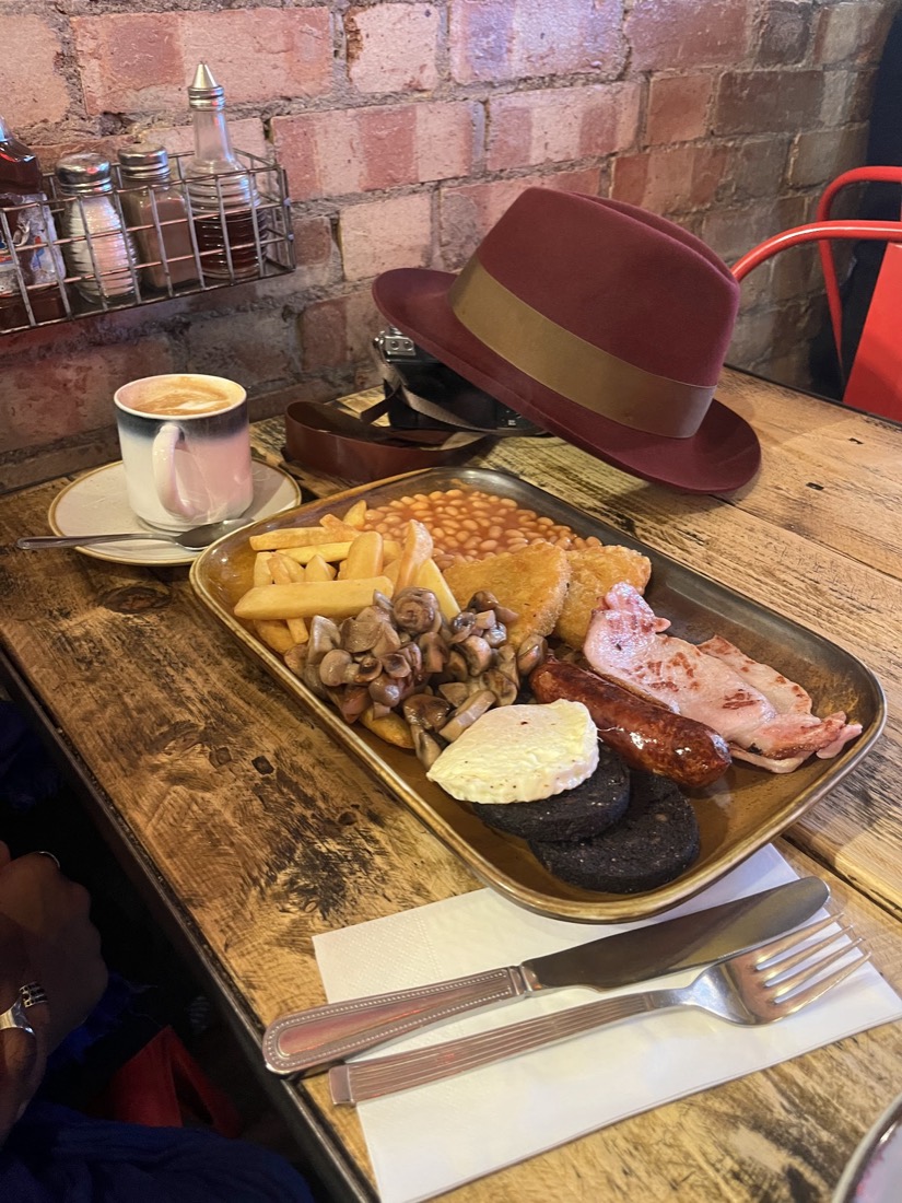 A full English breakfast on a metal tray at Nilly's Cafe: chips, beans, hash browns, mushrooms, sausage, bacon and black pudding, with a cappuccino against an exposed brick wall.