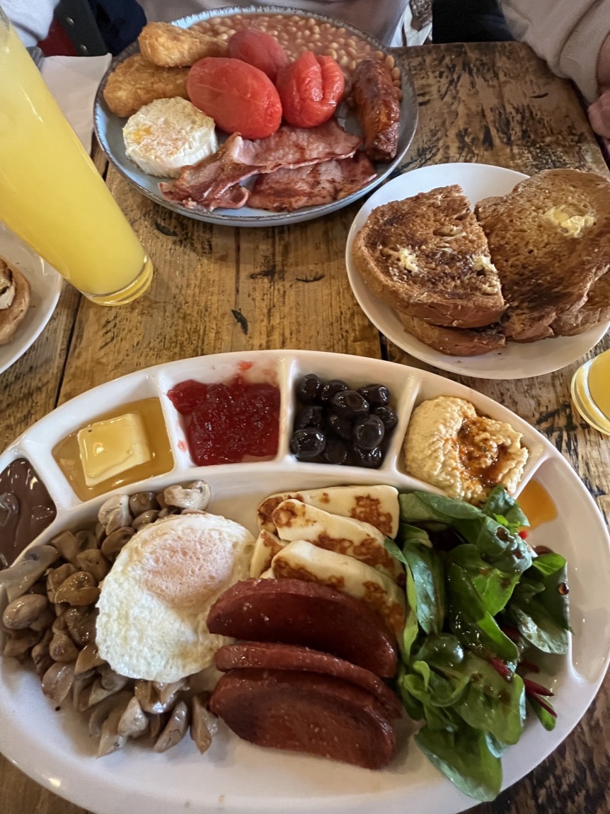 A generous Nilly's Cafe breakfast spread on a wooden table: full English with hash browns, beans and grilled tomatoes, a sharing platter with halloumi and hummus, toast and fresh orange juice.