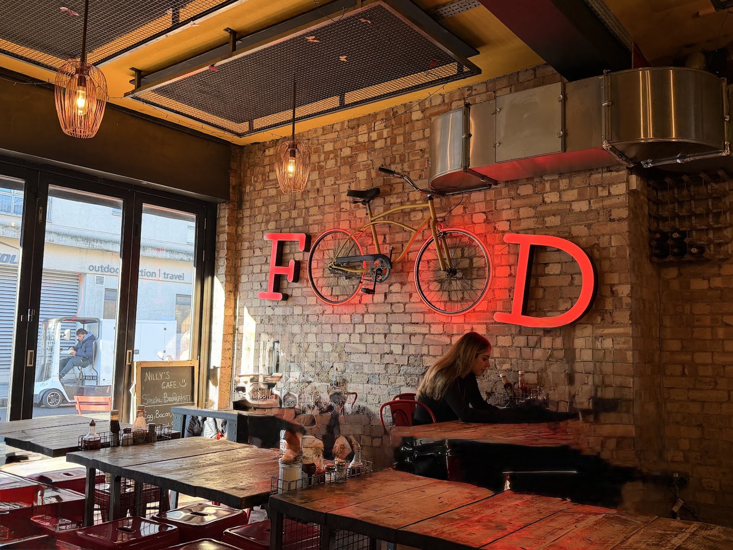 Inside Nilly's Cafe: exposed brick walls, reclaimed wood tables and red tolix chairs beneath a glowing red neon FOOD sign with a bicycle mounted on the wall.