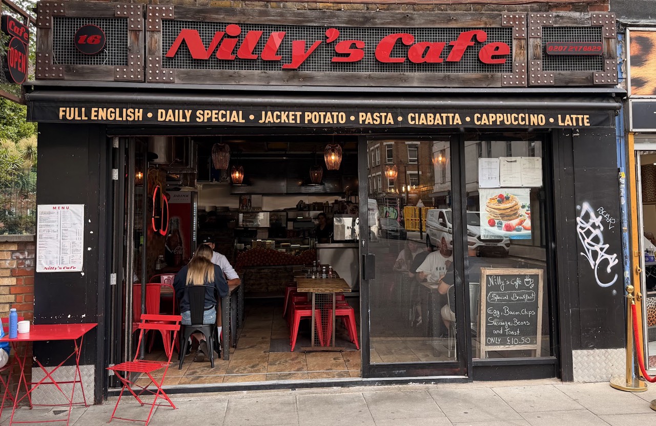 The Nilly's Cafe shopfront at 16 Bell Lane, a bold red sign on a black riveted timber facade reading Full English, Daily Special, Jacket Potato, Pasta, Ciabatta, Cappuccino, Latte, with red pavement tables.